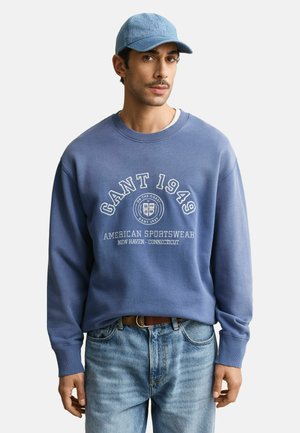 Homme portant un sweat-shirt bleu GANT 1949, une casquette en jean, un jean bleu clair, debout devant un fond blanc.