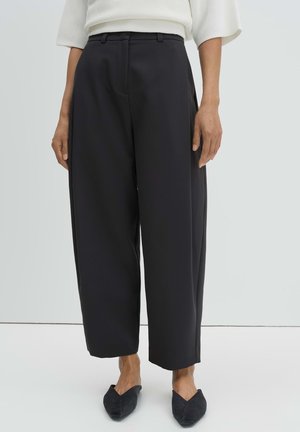 Persona che indossa pantaloni neri a vita alta e gamba larga abbinati a ballerine nere con punta appuntita e una maglietta bianca su uno sfondo uniforme.