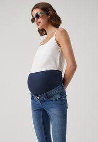 Graviditetsjeans med et marineblått elastisk panel, lyseblå denim og en hvit tanktop. Buksene har en klassisk passform med lukking med knapp.
