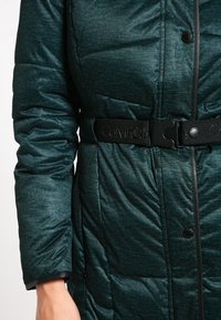 Tyrkysová prošívaná puffer bunda s páskem v pase, patentními knoflíky a texturovaným vzorem s černými akcenty.