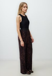 Schwarzes ärmelloses Oberteil kombiniert mit weiten, gemusterten Hosen in dunklen Braun- und Bronzetönen, die ein strukturiertes Design und einen hohen Bund aufweisen.