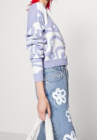 Lilac knit sweater with white floral pattern. Features ribbed cuffs and hem. Paired with blue jeans embellished with white flower designs.