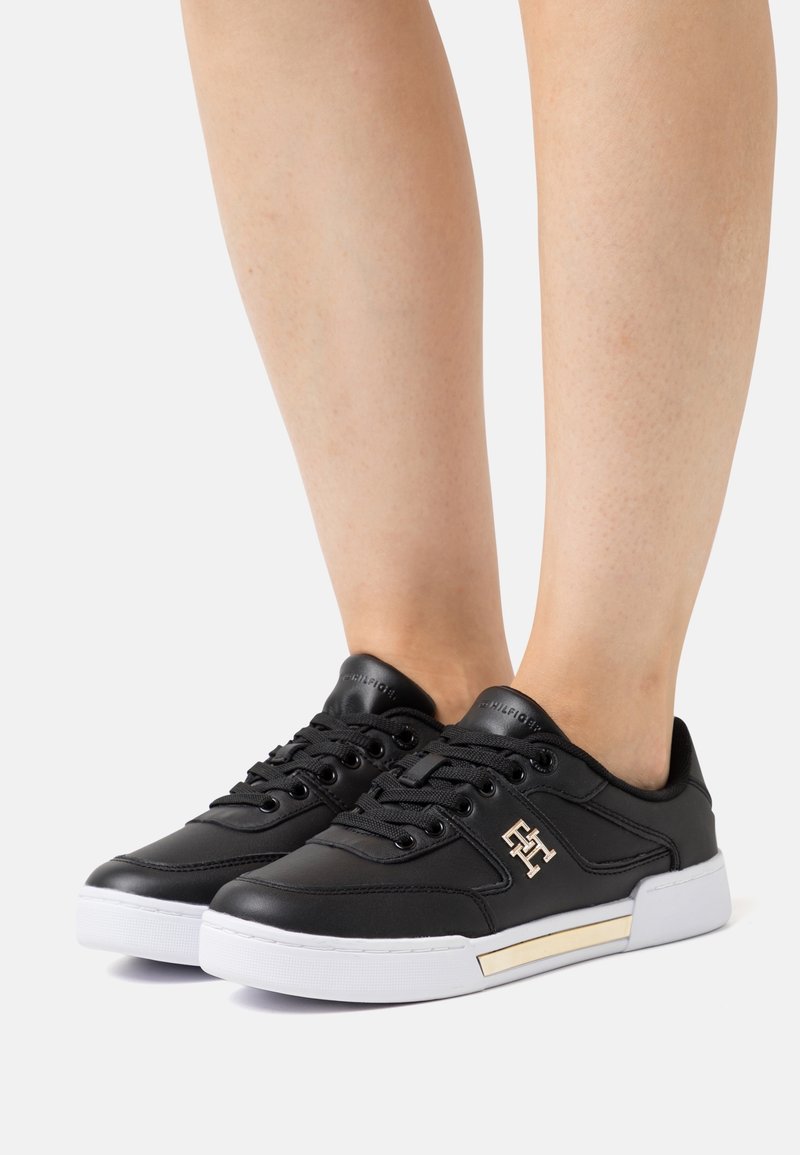 Baskets basses noires avec semelles blanches et détail du logo doré portées aux pieds, présentées sur un fond clair uni.