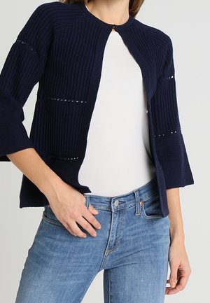 Cardigan en tricot bleu marine avec une texture côtelée, à l'avant ouvert, et des manches évasées avec des détails découpés. Associé à un haut blanc et un jean clair.