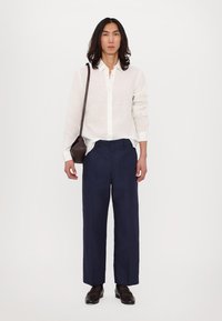 Fehér, hosszú ujjú lening póló, sötétkék személyre szabott nadrág, barna bőrcipő, és egy kis keresztpántos táska. A modell középen áll egy sima háttér előtt.