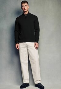 Crna pletena vesta s polu-zip ovratnikom, uparena s bijelim ravnim hlačama i crnim nekupljenim čizmama. Jednostavna pozadina.