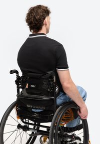 Άνδρας με σγουρά μαλλιά, μαύρο πουκάμισο και τζιν κινεί χειροκίνητο αναπηρικό καροτσάκι, από πίσω σε απλό λευκό φόντο.