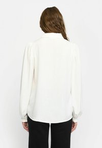 Weiße Bluse mit langen, gepufften Ärmeln, einem Kragen, einem leicht geschwungenen Saum und einer glatten Stoffstruktur, kombiniert mit schwarzen Hosen.