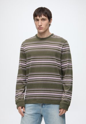 Giovane uomo che indossa una camicia a maniche lunghe verde oliva con sottili righe orizzontali bianche e rosa e jeans azzurri, in piedi davanti a uno sfondo uniforme.