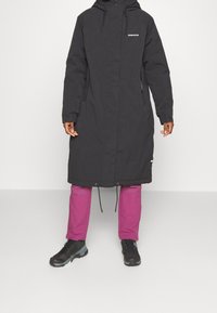 Cappotto lungo nero con cappuccio, dotato di chiusura a cerniera e lacci elastici. Abbinato a pantaloni rosa vivaci e scarpe nere robuste.