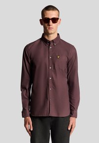 Camicetta bordeaux a maniche lunghe con colletto alla francese, taschino frontale e piccolo logo giallo. Indossata con occhiali da sole scuri e pantaloni neri con texture.