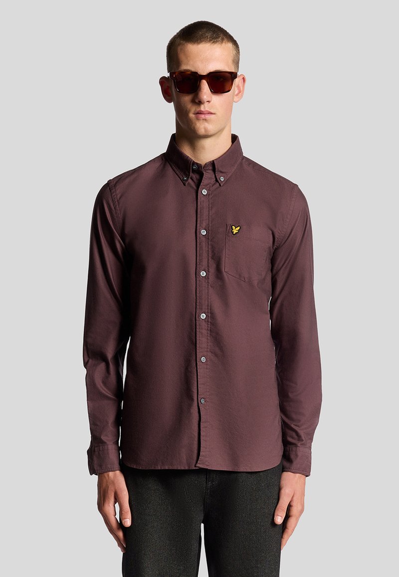 Camicetta bordeaux a maniche lunghe con colletto alla francese, taschino frontale e piccolo logo giallo. Indossata con occhiali da sole scuri e pantaloni neri con texture.