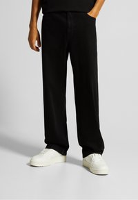 Pantalones de mezclilla negros de pierna recta, con un bolsillo frontal y combinados con zapatillas blancas, mostrando una textura suave y mate.