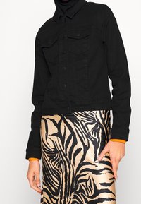 Femme portant une veste noire boutonnée sur une jupe beige et noire imprimée zèbre, avec un poignet orange visible au niveau du poignet.