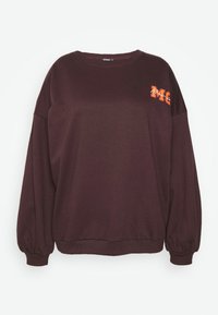 Felpa marrone scuro con scollatura rotonda, spalle cadute, polsini arricciati e un logo arancione "M" sul petto. Tessuto morbido.