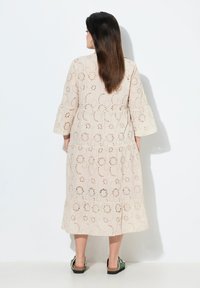 Cremfarbendes, besticktes Midi-Kleid mit floralen Ausschnitten und weit ausgestellten Ärmeln. Geschichtetes Design, strukturierter Stoff und geraffte Taille.