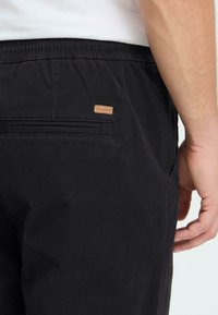 Un pantalon en coton noir avec une ceinture élastique et une poche arrière ornée d'une petite étiquette en cuir marron, présentant une texture lisse et un design décontracté.