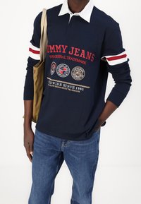 Navy long-sleeve polo shirt with white collar, red accents, and embroidered Tommy Jeans logo; worn with blue jeans and a tan tote bag.