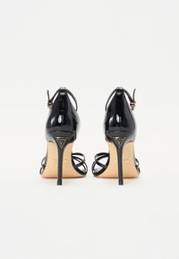 Sandalias de tacón alto de charol negro con parte trasera abierta, correas finas y un detalle de logo triangular en el tacón. Acentos de herrajes en tono dorado.
