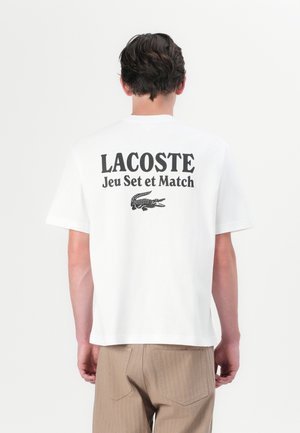 Homme portant un t-shirt blanc Lacoste avec un texte noir et le logo crocodile au dos, accompagné d'un pantalon marron clair, se tenant devant un fond uni.