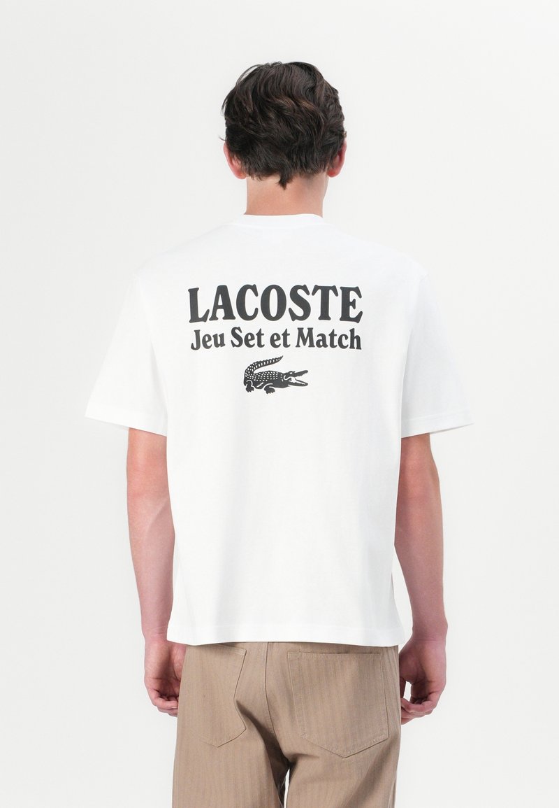 Homme portant un t-shirt blanc Lacoste avec un texte noir et le logo crocodile au dos, accompagné d'un pantalon marron clair, se tenant devant un fond uni.