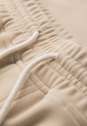 Beige elastische tailleband met witte trekkoord op gestructureerde stof, waarschijnlijk onderdeel van casual of sportbroeken of shorts.