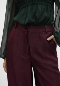 Burgunderfargede plissert bukser med en glat tekstur, som har en knappelukking og sidelommer, sammen med en transparent grøn bluse.