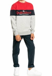 Sudadera gris con rayas horizontales rojas y marinas, con el logotipo de "Quiksilver". Combinada con pantalones marinos y zapatillas blancas.