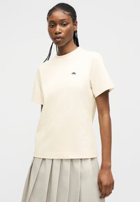 Νεαρή γυναίκα με πλεξούδες, ντυμένη με απλό μπεζ κοντομάνικο μπλουζάκι και ανοιχτό μπεζ πλισέ φούστα, που στέκεται μπροστά σε ένα απλό φόντο.