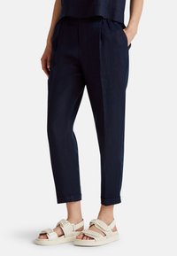 Navy linen trousers with side pockets, tapered leg, and raw hem. Worn with white open-toe sandals featuring a woven strap design.