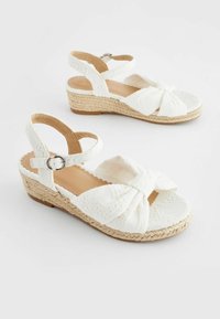 Sandali con tacco bianco a forma di cuneo con un design tessuto e lavorato. Presentano un grande fiocco sulla parte anteriore, una cinghia alla caviglia con una fibbia argentata e una soletta avvolta in juta.