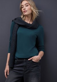 Mørkegrønt langærmet top med rund halsudskæring, parret med mørkegrå jeans. Begge dele er suppleret af et mørkegrå tørklæde med en blød tekstur.