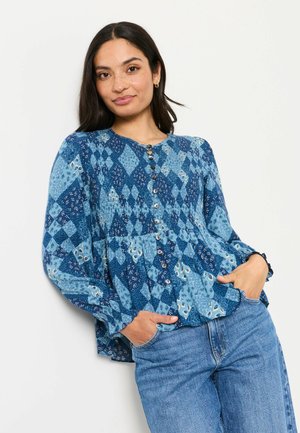 Frau trägt eine blau gemusterte langärmlige Bluse mit Knöpfen und hellblaue Jeans, steht mit den Händen in den Taschen vor weißem Hintergrund.