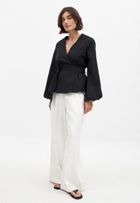 Blusa negra con mangas largas y voluminosas, ceñida a la cintura con un cinturón, combinada con pantalones anchos blancos y sandalias negras.