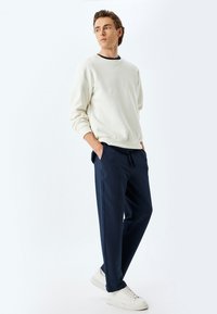 Sudadera beige clara con cuello redondo; pantalones deportivos texturizados en navy con cintura elástica; zapatillas blancas; de pie contra un fondo plano.