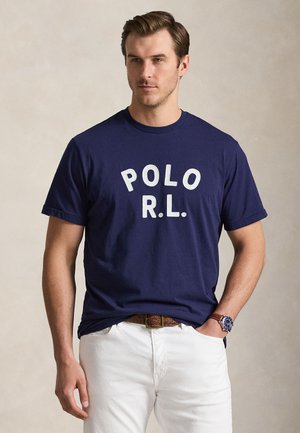 Homme portant un t-shirt bleu marine avec le texte "POLO R.L.", un pantalon blanc, une ceinture marron et une montre bleue au poignet, debout avec une main dans la poche.