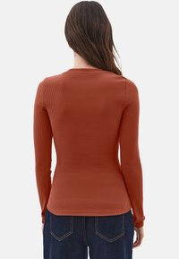 Mujer con cabello largo castaño que lleva una camiseta ajustada de manga larga color óxido y jeans oscuros, de espaldas a la cámara.