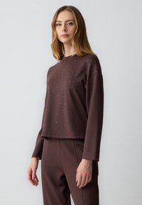 Maglietta a maniche lunghe marrone con vestibilità rilassata, decorata con piccoli chiodi metallici disposti in modo irregolare sul davanti.