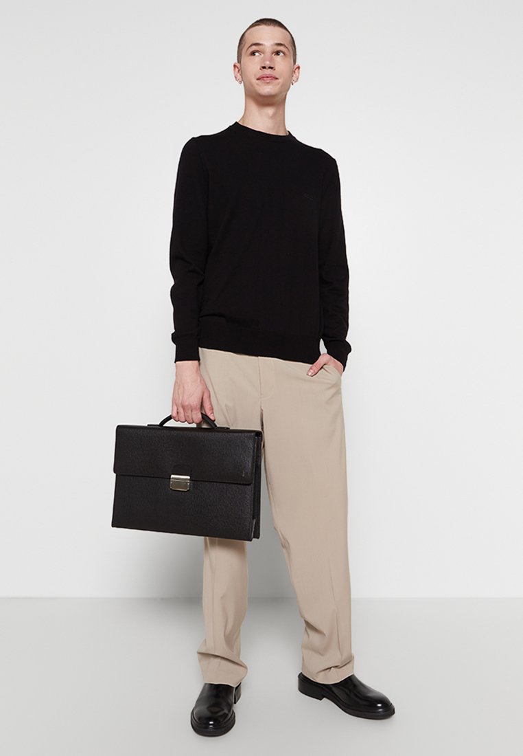 Zwarte trui, beige broek en zwarte leren schoenen. Draagt een gestructureerde zwarte aktetas met een metalen sluiting. Eenvoudig, modern ontwerp.