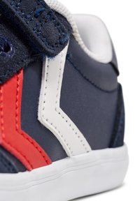 Marineblauwe sneaker met rode en witte zigzagaccenten, klittenbandsluiting, gestructureerd bovenmateriaal en een witte rubberen zool. Close-up detail.