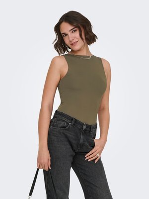 Kobieta o krótkich falowanych włosach, ubrana w oliwkowy top bez rękawów i czarne dżinsy, trzymająca czarną torebkę na pasku prawą ręką.