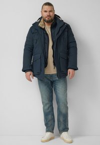 Granatowa parka z kapturem, z teksturowaną podszewką wewnętrzną, suwakiem z przodu, dwoma przednimi kieszeniami i regulowanymi mankietami, zestawiona z jasnoniebieskimi dżinsami i białymi trampkami.