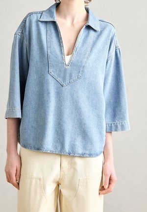 Lichtblauw denim shirt met een kraag, diepe V-hals en driekwart mouwen. Heeft onafgewerkte zomen en een losse pasvorm. Gebruikt met een beige broek.