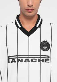 Jeune personne portant une chemise blanche avec des rayures verticales noires, un col noir, et le texte "PANACHE" ainsi qu'un logo rond sur la poitrine.