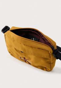 Žltá nylonová taška cez rameno so zipsovým otvorom a vnútornou podšívkou v bordovej farbe. Má predné vrecko s detailom v podobe loga Calvin Klein.