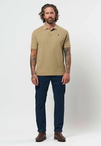 Homme aux cheveux bouclés et tatouages portant un polo beige, un pantalon bleu marine et des bottes marron, debout devant un fond clair uni.