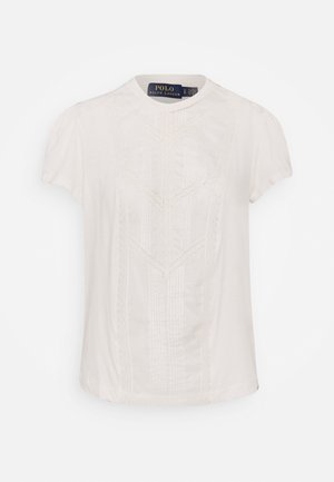 Biała bluzka z krótkim rękawem z delikatnymi koronkowymi zdobieniami i plisowanym wzorem, okrągłym dekoltem oraz metką Polo Ralph Lauren wewnątrz kołnierza.