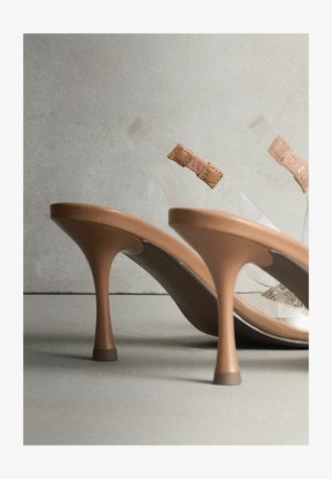 Bőrszínű magas sarkú szandálok sima felülettel, átlátszó pántokkal és texturált díszítéssel a bokánál. Elegáns, keskeny tűsarkú kialakítás.