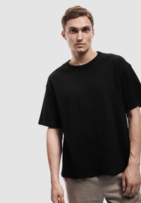 Ung mand med kort brunt hår iført en ensfarvet sort T-shirt med korte ærmer og beige bukser mod en lys grå baggrund.