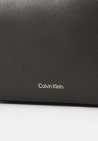 Juodas odinis piniginis su tekstūruota paviršiumi, su auksiniu "Calvin Klein" logotipu išpjaustytu priekyje, suteikiančiu papildomą detalumą.